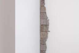 A gray, vertical stack of bricks with one straight edge and one jagged edge is displayed against a white wall in a gallery corner.