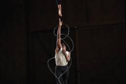 A dancer in a wheelchair is suspended from the ceiling. Her right arm clasps a cable that’s hooked onto her wheelchair, and her left reaches down toward another dancer, who jumps and extends his right arm toward her; his left arm rests on his chest. He is encircled by silver barbed wire.